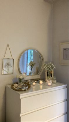 a white dresser with flowers and candles on it in front of a round mirror above the dresser