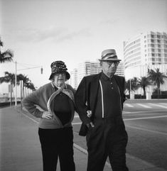 an older man and woman walking down the street together, with palm trees in the background