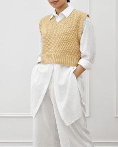 a woman standing in front of a white wall wearing a yellow sweater and white pants