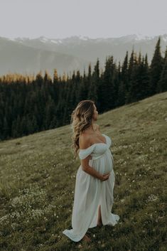 a pregnant woman standing on top of a grass covered field next to trees and mountains