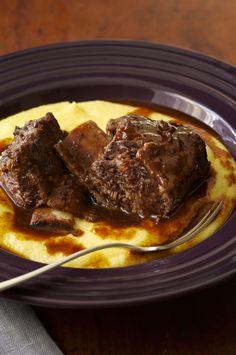 a purple plate topped with meat and grits covered in gravy next to a fork