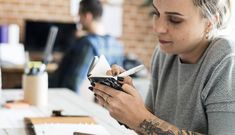 a woman sitting at a table looking at her cell phone while holding a pen and paper