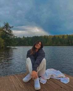 a woman sitting on top of a wooden dock next to a body of water with trees in the background