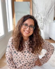 a woman with glasses is sitting on a bed in front of a window and smiling at the camera