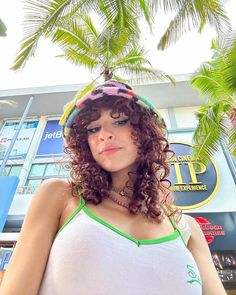 a woman with curly hair wearing a green and white tank top standing in front of a palm tree