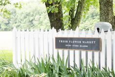 a white picket fence with a sign that reads fresh flower market next to trees and flowers