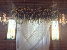 a white drape with flowers and greenery hanging from it's sides in front of two windows