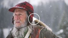 an old man wearing a red and black hat in the snow with trees behind him