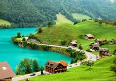 a lake surrounded by lush green hills and houses next to it with mountains in the background