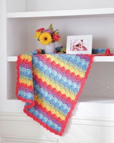 a crocheted blanket on a shelf with flowers and pictures in the back ground