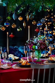 the table is set for christmas dinner with candles and ornaments hanging from the tree in the background