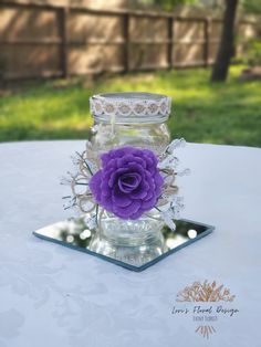 a purple flower sitting in a glass jar on top of a white cloth covered table