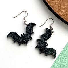 Midnight Flight: Black Glitter Bat Dangle Earrings! Take flight with these striking black glitter bat earrings, perfect for adding a touch of mystery to any outfit. Crafted from shimmering black glitter acrylic, these bat-shaped earrings sparkle with every move, creating an eye-catching accessory for Halloween or any time you want to embrace your dark side. Measuring approximately 1.5 inches from wing to wing, they're the perfect size for a bold yet lightweight statement piece. - **Material Black glitter acrylic   - **Size Approximately 1.5 inches from wing to wing   - **Earring Style Dangle earrings with stainless steel wire hooks   - **Lightweight & Comfortable** for all-day wear   - Great for Halloween, gothic fashion, or year-round spooky vibes! Whether you're dressing up for a Hallowe Embrace Your Dark Side, Flying Bat, Bat Earrings, Halloween Gothic, Glitter Acrylic, Glitter Earrings, Glitter Acrylics, Spooky Vibes, Sparkle Earrings