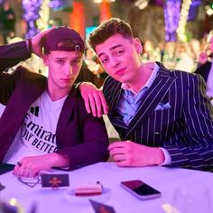 two young men sitting at a table with cell phones in front of them, one is wearing a hat and the other has his arm around his neck