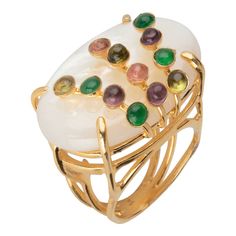 The Giselle Ring features a substantial oval mother of pearl stone adorned with tourmaline small stones, creating a vintage and art deco-inspired style.  Stones: Mother of Pearl & Tourmaline  Material: 14K Gold Plated  Dimensions: 1 1/4 X 7/8" Luxury White Oval Moonstone Ring, Luxury White Multi-stone Gemstones, Luxury White Opal Cabochon Ring, Luxury White Oval Cabochon Ring, Luxury Oval Rings With Natural Stones, Elegant Multi-stone Opal Ring With Oval Cabochon, Oval Multi-stone Moonstone Ring For Formal Occasions, Formal Oval Multi-stone Moonstone Ring, Art Deco Oval Opal Ring