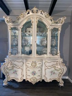 an ornate china cabinet with glass doors and drawers on wheels, painted in antique white