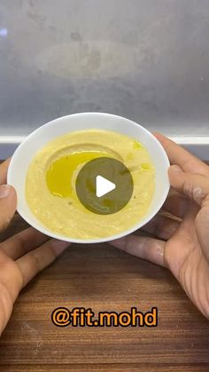 two hands holding a white bowl filled with yellow liquid and an olive in the middle