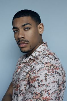 a man with a mustache and flowered shirt looks at the camera while standing in front of a blue background