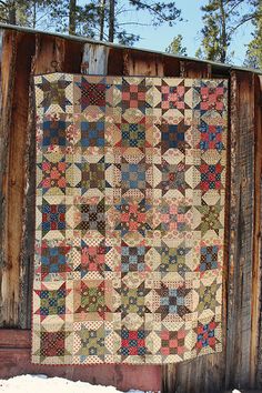 an old quilt hanging on the side of a wooden building with snow around it and trees in the background