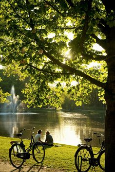 two people sitting on a bench next to bicycles near a lake with a fountain in the background