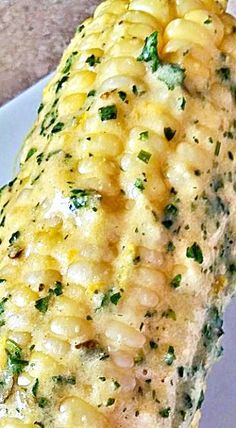 a corn on the cob with cheese and green herbs is sitting on a white plate