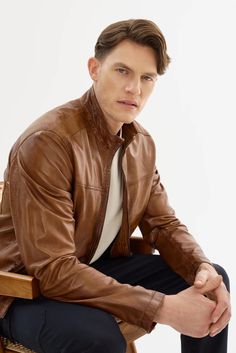 a man in a brown leather jacket sitting on a wooden chair with his hands crossed