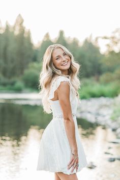 a woman standing in front of a body of water wearing a white dress and smiling at the camera