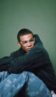 a young man wearing glasses sitting on the floor with his arms crossed and looking at the camera