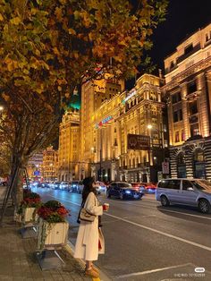 a woman standing on the side of a road next to a tree and buildings at night