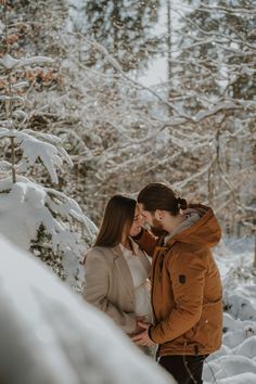 a man and woman standing in the snow near some trees with their faces close to each other