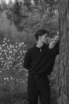 a man standing next to a tree in a forest with wildflowers behind him