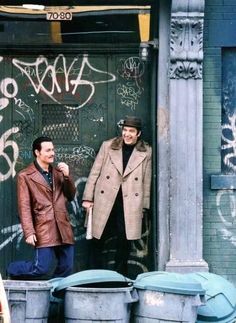 two men standing in front of a door with graffiti on the wall and trash cans