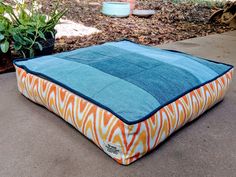 an orange, blue and white dog bed sitting on the ground next to some potted plants