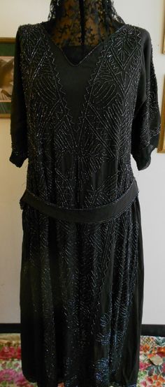 "1920's, 38\" bust, light weight black silk crepe gown entirely sewn in iridescent black glass beaded Art Deco abstract geometric design, from shoulder to hem. A matching sash edged in the same beads is attached to the gown at upper hip. Neckline of gown is open to a small V, with a modesty of black chiffon at the bottom of the V, neckline is edged with a single row of iridescent beads. The gown has short raglan sleeves, covered with the same beaded motif, edged in a narrow band of chiffon. Bodi Black Art Deco Flapper Dress With Sequins, Black Sequined Art Deco Flapper Dress, Black Gatsby Style Embellished Dress, Black Art Deco Flapper Dress For Evening, Vintage Black Flapper Dress With Beaded Fringe, 1920s Beaded Evening Dress, 1920s Beaded Fringe Evening Dresses, 1920s Style Beaded Fringe Evening Dresses, 1920s Evening Dresses With Beaded Fringe