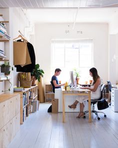 two people sitting at a desk in an office with boxes on the shelves and bookshelves