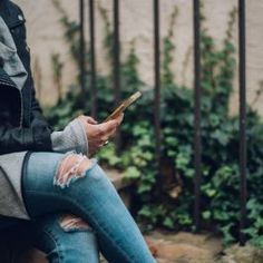 a person sitting on a bench looking at their cell phone while wearing ripped jeans and a leather jacket