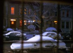 snow covered cars are parked in front of a building at night, seen through a window