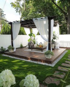 a wooden deck surrounded by white curtains and potted plants in the middle of a yard