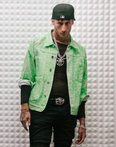 a young man wearing a green jacket and black hat standing in front of a wall