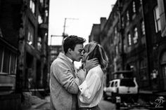 a man and woman kissing on the street in front of some old buildings with cars behind them