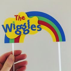 a hand holding up a sign that says the wiggles on it with a rainbow in the background