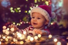 a baby wearing a santa hat sitting in front of a christmas tree filled with lights