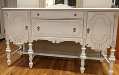 an antique white painted buffet table in a room with hardwood floors and walls, along with a mirror on the wall