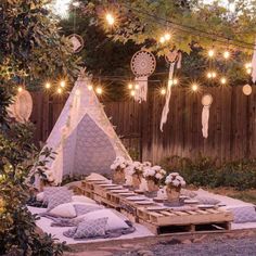 an outdoor dining area with lights strung over the table and pillows on the ground, in front of a wooden fence