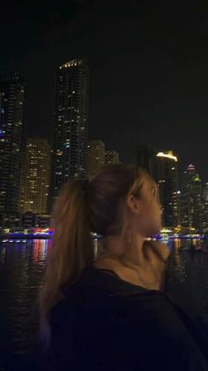 a woman standing in front of a city skyline at night with lights reflecting off the water