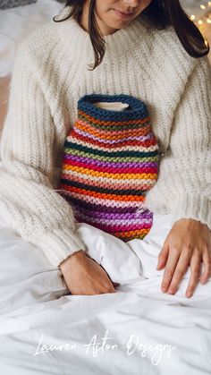 a woman sitting on top of a bed wearing a sweater and holding a knitted purse