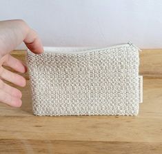 a hand is holding onto a white knitted pouch on a wooden table with a white wall in the background