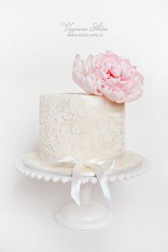 a white wedding cake with pink flower on top and ribbon around the edge, sitting on a pedestal