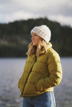 a woman in a yellow jacket and white hat standing next to the water with her hands on her hips