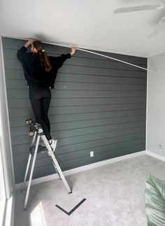 a woman standing on a stepladder in front of a gray wall and ceiling fan